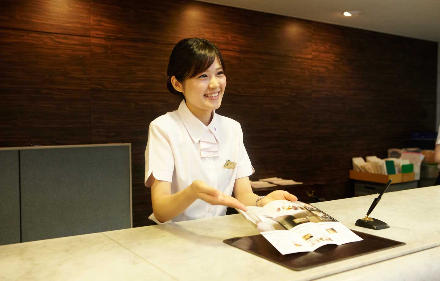 爽やかで素敵な笑顔でホテルパンフレットを紹介するホテルフロントスタッフ
